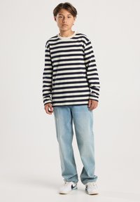 Junge Person steht, trägt ein schwarz-weiß gestreiftes Langarmshirt, hellblaue Jeans und weiße Sneakers mit schwarzem Swoosh.