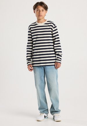 Jongere staat rechtop, gekleed in een zwart-witte gestreepte lange mouwen shirt, lichtblauwe spijkerbroek en witte sneakers met zwarte swoosh.