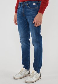 Uomo che indossa jeans di denim blu con etichetta Roy Rogers e sneakers bianche con accenti beige e neri, in piedi contro uno sfondo semplice.