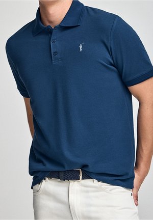 Mies, joka pukeutunut tummansiniseen lyhythihaiseen poolopaitaan, jossa on pieni valkoinen golfarin logo, valkoisiin housuihin ja tummansiniseen kudottuun vyöhön.