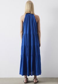 Robe maxi bleue, à volants, avec un décolleté halter. Le tissu semble léger et texturé, tombant juste au-dessus des chevilles. Accessoires minimalistes.