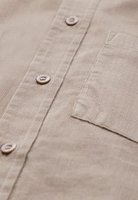 Camicia di ciniglia beige con costine verticali, caratterizzata da bottoni argentati e una tasca sul petto. Il tessuto ha una texture morbida e una lucentezza sottile.