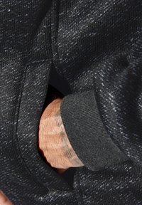 Black textured fabric with a flecked pattern; a hand is inserted into a side pocket. Visible cuff in a contrasting solid black material.