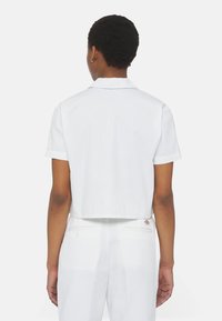 Camicia bianca cropped con maniche corte e colletto classico, caratterizzata da una texture liscia e un discreto logo sul retro.