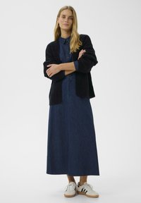 Dunkelblaues Denimkleid mit einer Knopfleiste vorne und einem langen A-Linien-Schnitt, kombiniert mit einer schwarzen Strickjacke und weißen Sneakers mit schwarzen Streifen.