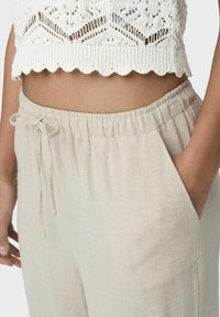 Pantalon en lin beige clair à coupe décontractée, muni d'une taille élastique, d'un cordon de serrage et de poches latérales. La texture semble légèrement froissée.