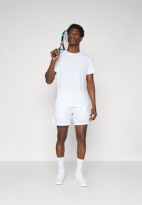 Hvite sportsklær bestående av en kortermet skjorte og shorts, kombinert med hvite sokker og sko, og holder en blå og svart tennisracket.