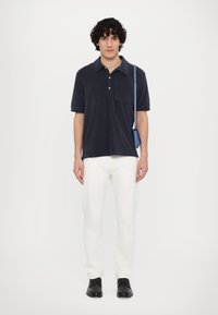 Marinblå polo t-shirt med korta ärmar och ficka, kombinerad med vita jeans. Modellen bär en liten blå axelväska och har på sig svarta skor.