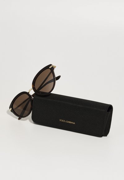 Schwarze Sonnenbrille im Cat-Eye-Design, mit dunkelbraunen Gläsern und goldenen Akzenten am Rahmen, die auf einem schwarzen Etui mit goldenem Logo ruhen.