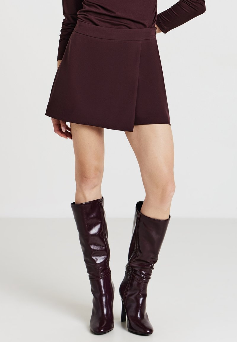 Donna con una mini gonna viola scuro e stivali in pelle lucidi con tacco alti fino al ginocchio su uno sfondo bianco semplice.