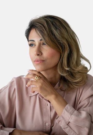 Goldring mit geflochtenem Design, am rechten Handgelenk getragen. Hellrosanes Hemd mit Knöpfen, hochgeklappten Ärmeln und glatter Textur. Braune, gestylte Haare.