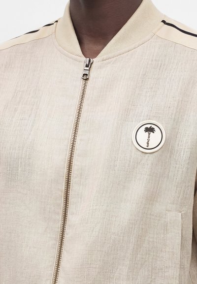 Ανοιχτό μπεζ ζακέτα με φερμουάρ, ριμπ γιακά, μαύρη ρίγα στον ώμο και στρογγυλή μπαλώματα με φοίνικα και το κείμενο "Palm Angels".