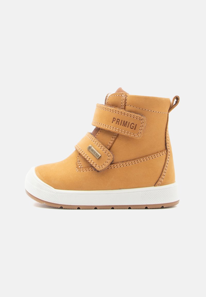 Bottines en cuir tanné avec deux sangles Velcro, semelle en caoutchouc blanche et accents cousus. Présente une boucle à l'arrière pour un enfilage facile.