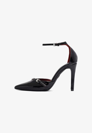 Black patent leather high-heeled shoe with pointed toe, ankle strap, and silver hardware. Glossy finish; red interior. 10 cm heel height.