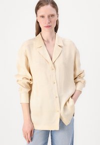 Lys beige skjorte med knapper laget av et teksturert stoff, med krage, lange ermer og subtile bladmønstre. Lommer på sidene.
