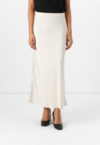 Maxi skirt satin krim dengan tekstur halus, gesper samping, dan pinggang yang pas. Dipadukan dengan atasan hitam dan sepatu hak tinggi hitam.