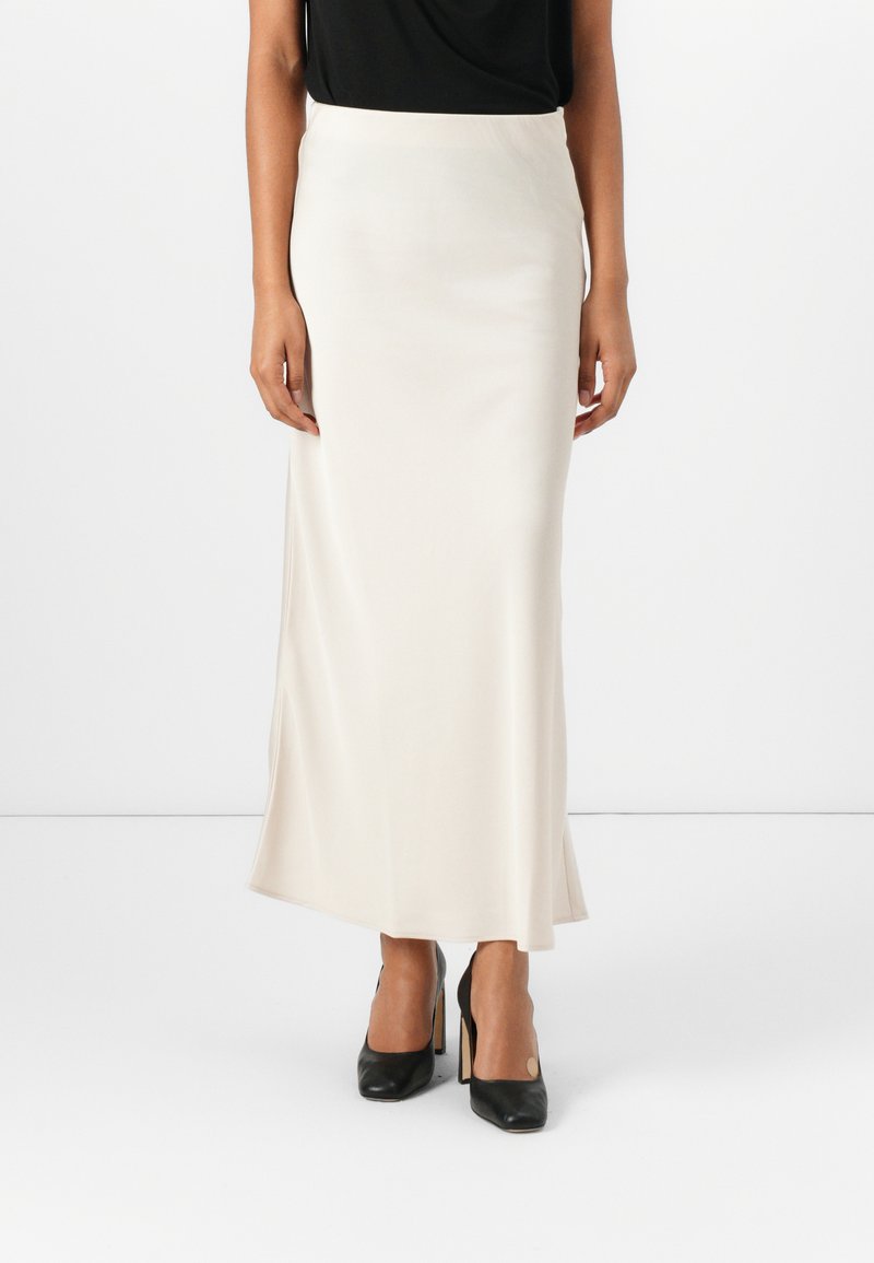 Maxi skirt satin krim dengan tekstur halus, gesper samping, dan pinggang yang pas. Dipadukan dengan atasan hitam dan sepatu hak tinggi hitam.