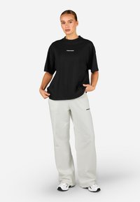 Melnā kokvilnas T-krekls ar vaļīgu piegriezumu un īsām piedurknēm, kombinēts ar gaiši pelēkām plaša piegriezuma sporta biksēm ar logotipu uz kreisās kājas.