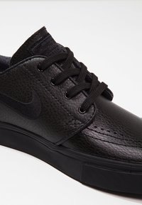 Svart lädersneaker med en texturerad yta, platta snören och en gummisula. Anmärkningsvärd Nike-logotyp på sidan och minimala sömnadsdetaljer.