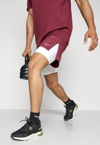 Shorts de sport bordeaux avec des accents blancs, chaussures de sport noires et un t-shirt bordeaux assorti, tenant un kettlebell noir.