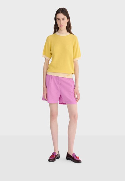 Femme aux longs cheveux bruns portant un pull jaune à manches courtes, un short rose et des mocassins bicolores violets et bordeaux avec des pampilles.