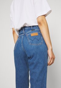 Světle modré džíny s vysokým pasem, viditelnými zadními kapsami a hnědou koženou nášivkou s nápisem "Wrangler" nad pravou kapsou.