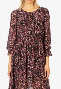 Robe florale en tissu noir transparent avec des fleurs roses et blanches. Présente des manches longues bouffantes, un décolleté en trou de serrure et une taille froncée.