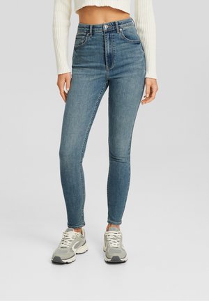 Femme portant un jean skinny bleu taille haute, un pull court blanc côtelé et des baskets épaisses grises et blanches, debout sur un fond gris clair.