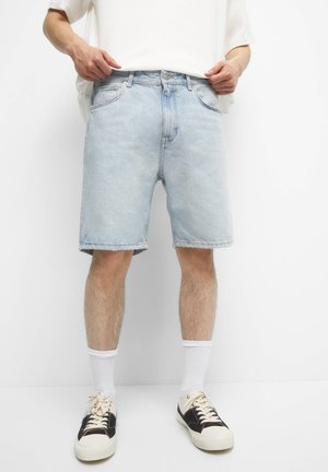 Personne portant un short en jean bleu clair, des chaussettes blanches, des baskets noires et blanches, ajustant sa chemise blanche.