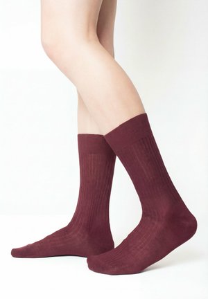 Beine mit dunkelroten, gerippten Socken bis zur Mitte der Wade, stehend auf weißem Hintergrund, wobei ein Fuß leicht angehoben ist.