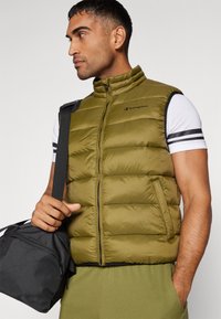 Olivgrüne Puffer-Weste mit hohem Kragen, Frontreißverschluss und Champion-Logo. Das Modell trägt ein weißes Hemd mit schwarzen Streifen und hält eine schwarze Tasche.