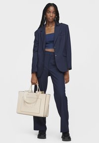 Costume bleu marine à rayures, comprenant un long blazer et un pantalon ajusté, associé à un haut court et un sac fourre-tout crème. Tissu lisse, design élégant.