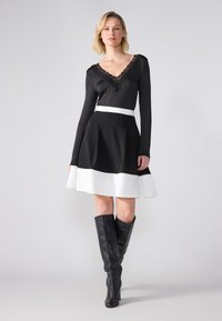 Robe noire à manches longues avec décolleté en V en dentelle, ceinture blanche, et ourlet évasé blanc. Portée avec des bottes noires jusqu'au genou. Texture lisse.