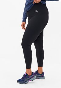 Schwarze Leggings aus elastischem Material mit glatter Textur, die ein dezentes Logo am Oberschenkel aufweisen und mit blauen Laufschuhen kombiniert werden.