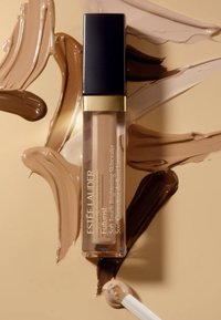 Estee Lauder Futurist Soft Touch Brightening Skincealer i en transparent tube med svart lokk, omringet av swatcher av beige og brune foundationnyanser.