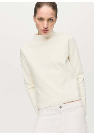 Mujer con el cabello recogido, vestida con un suéter crema de cuello alto y pantalones blancos, de pie frente a un fondo neutro.