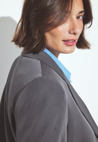 Jeune femme aux cheveux bruns courts portant un blazer gris et une chemise bleue, se retournant par-dessus son épaule contre un fond uni.