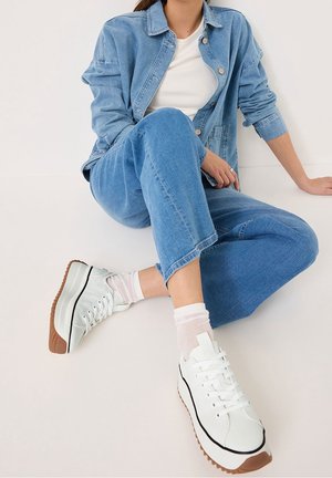 Osoba siedząca ze skrzyżowanymi nogami, ubrana w jasnoniebieską kurtkę jeansową i dżinsy, białe skarpetki oraz białe sneakersy na platformie z brązowymi podeszwami.