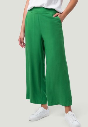 Vrouw met groene wijd uitlopende broek, witte sneakers en een wit topje, staand met één hand in de zak tegen een effen achtergrond.