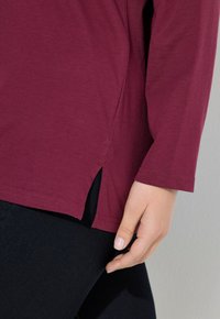 Bordeaux langpols blouse met zijsplitten, gemaakt van zachte katoenstof, gecombineerd met donkerblauwe jeans, wat een casual uitstraling geeft.