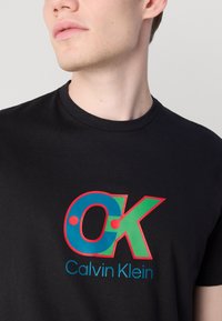 Juodas medvilninis marškinėlis su spalvingu "CK" logotipu su mėlynais, žaliais ir raudonais akcentais, o žemiau parašyta "Calvin Klein" mėlyna spalva.