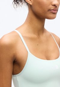 Νεαρή γυναίκα που φοράει ανοιχτοπράσινο τοπ με λεπτές τιράντες και το λογότυπο The North Face στο στήθος, εμφανιζόμενη από τον ώμο έως το πιγούνι.