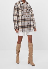 Kostkovaná bunda v béžové, hnědé a černé barvě s límcem a dvěma předními kapsami, kombinovaná s bílými šortkami a béžovými semišovými kozačkami sahajícími ke kolenům.