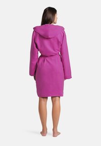 Accappatoio rosa con cappuccio realizzato in morbido tessuto peluche. Presenta una cintura da annodare, maniche lunghe e lunghezza fino al ginocchio. Senza motivi o elementi metallici.