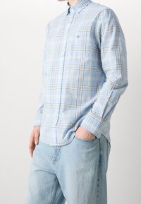 Camicia a quadri blu con bottoni in tessuto leggero, caratterizzata da linee bianche e verdi, taschino sul petto e colletto classico. Indossata con jeans chiari.