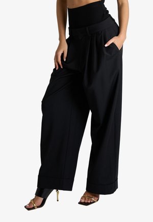 Donna che indossa pantaloni neri a vita alta e gamba larga, con le mani nelle tasche e sandali dorati con tacco alto e punta quadrata.