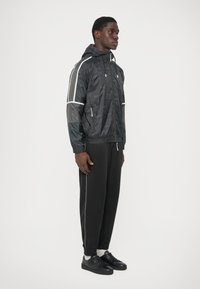 Vedere laterală a unui bărbat în picioare, purtând o jachetă cu glugă neagră cu dungi albe, pantaloni jogger negri, șosete albe și adidași negri.