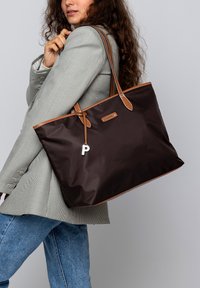 Bolso tote marrón hecho de nailon con acentos de cuero marrón claro. Presenta una prominente etiqueta con la letra "P" y costura en la parte superior a lo largo de los bordes.