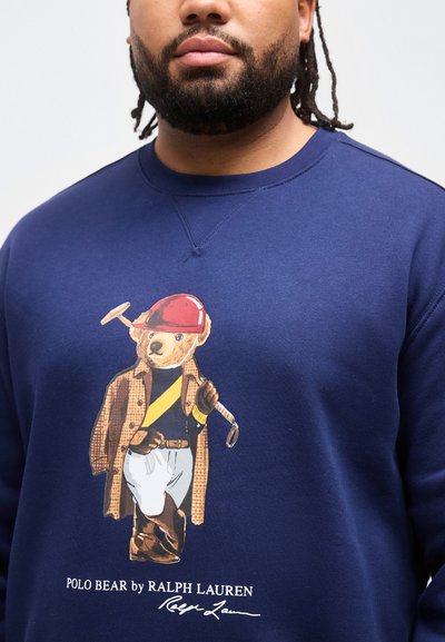 Άνδρας που φοράει μπλε σκούρο φούτερ με εικονογράφηση του Polo Bear ντυμένου με ιππικό εξοπλισμό, κρατώντας ρόπαλο πόλο, και το κεί μενο «Polo Bear by Ralph Lauren».