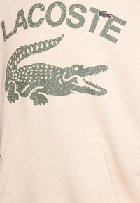 Felpa beige con un grande motivo di coccodrillo verde screpolato e la scritta "LACOSTE" sopra. Presenta una tasca a marsupio sul davanti.